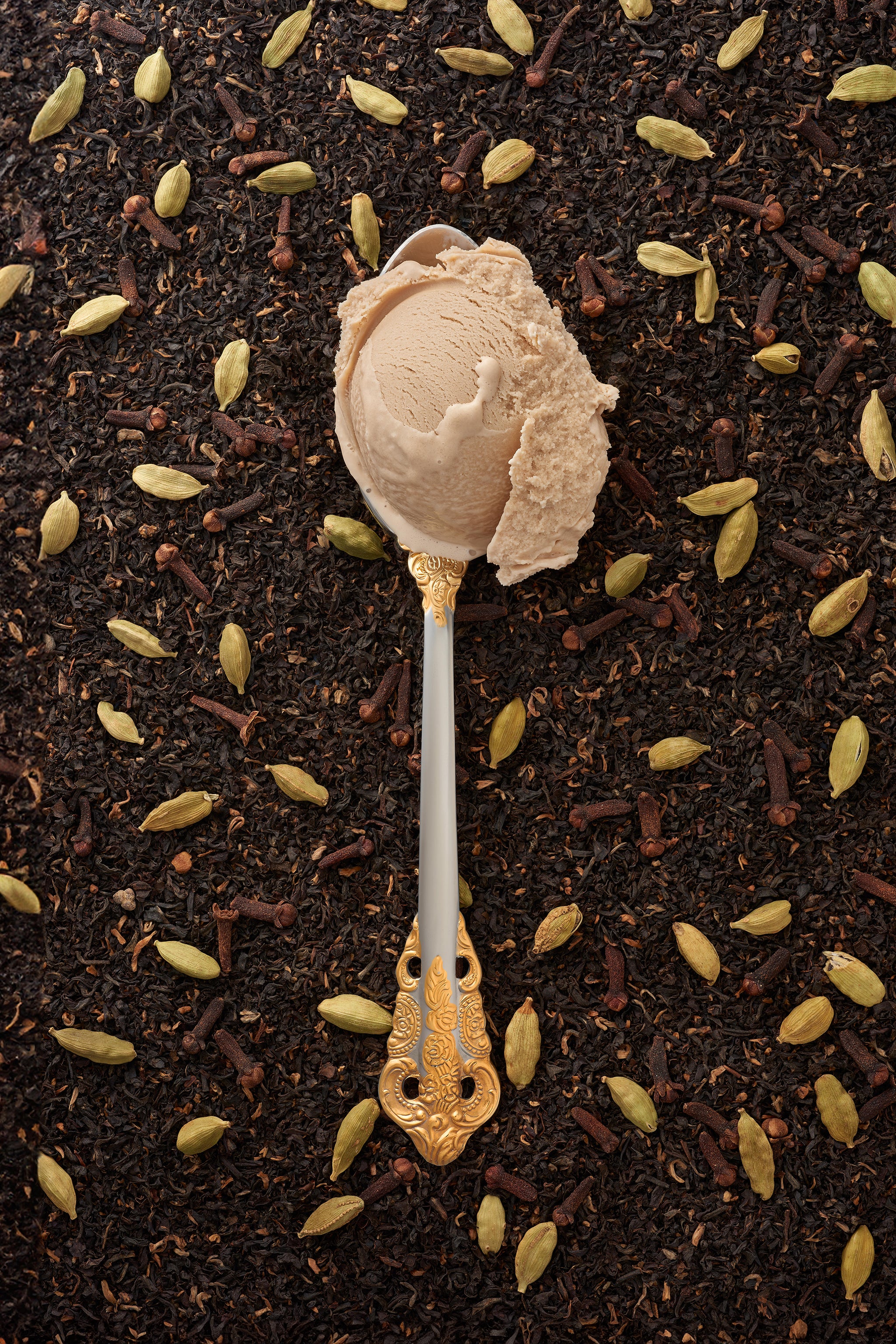 A scoop of cardamom-chai kulfi in a silver spoon with a gold handle, with a scattering of chai tea and cardamom pods in the background.