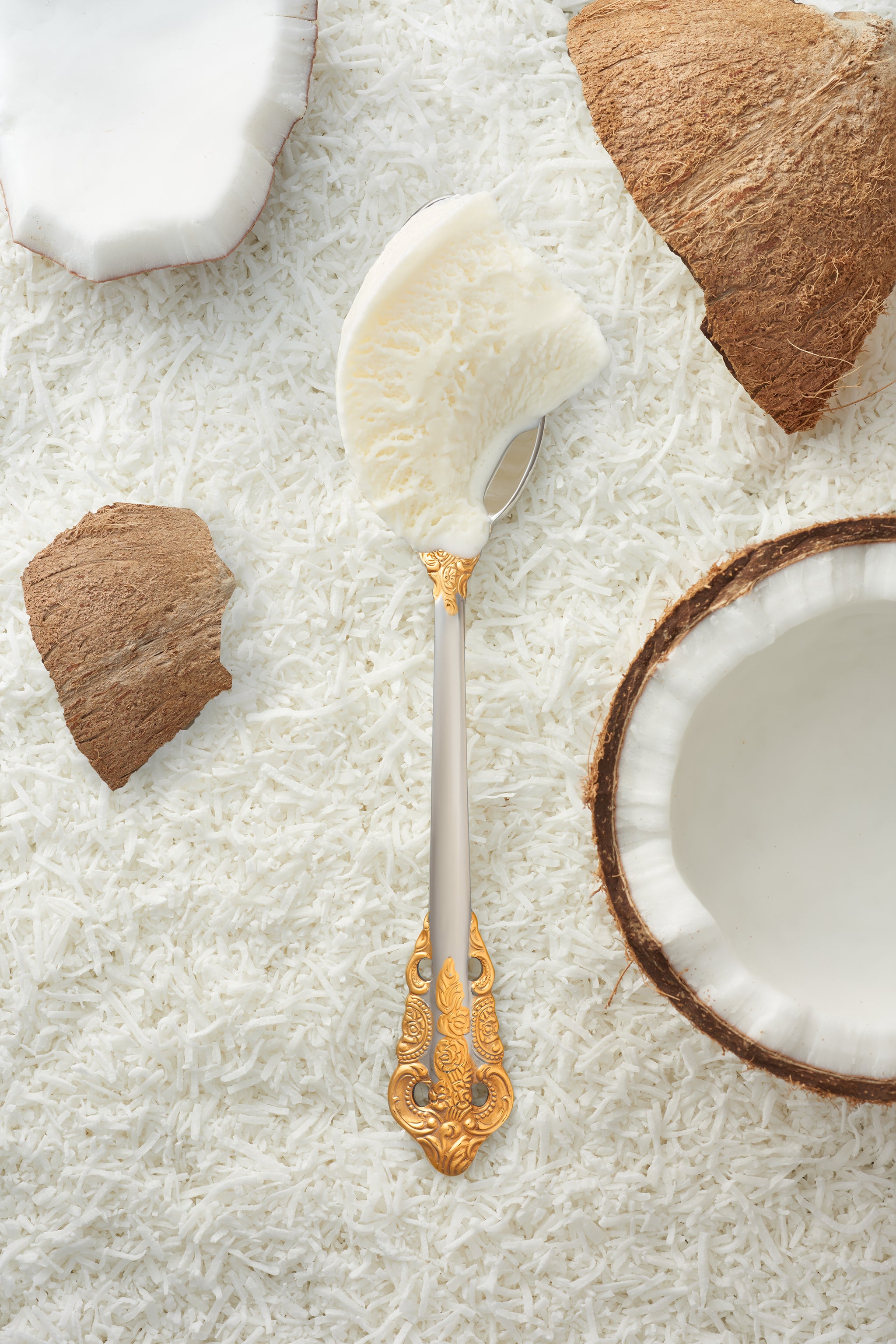 A scoop of coconut kulfi in a silver spoon with a gold handle, surrounded by pieces of fresh coconut.