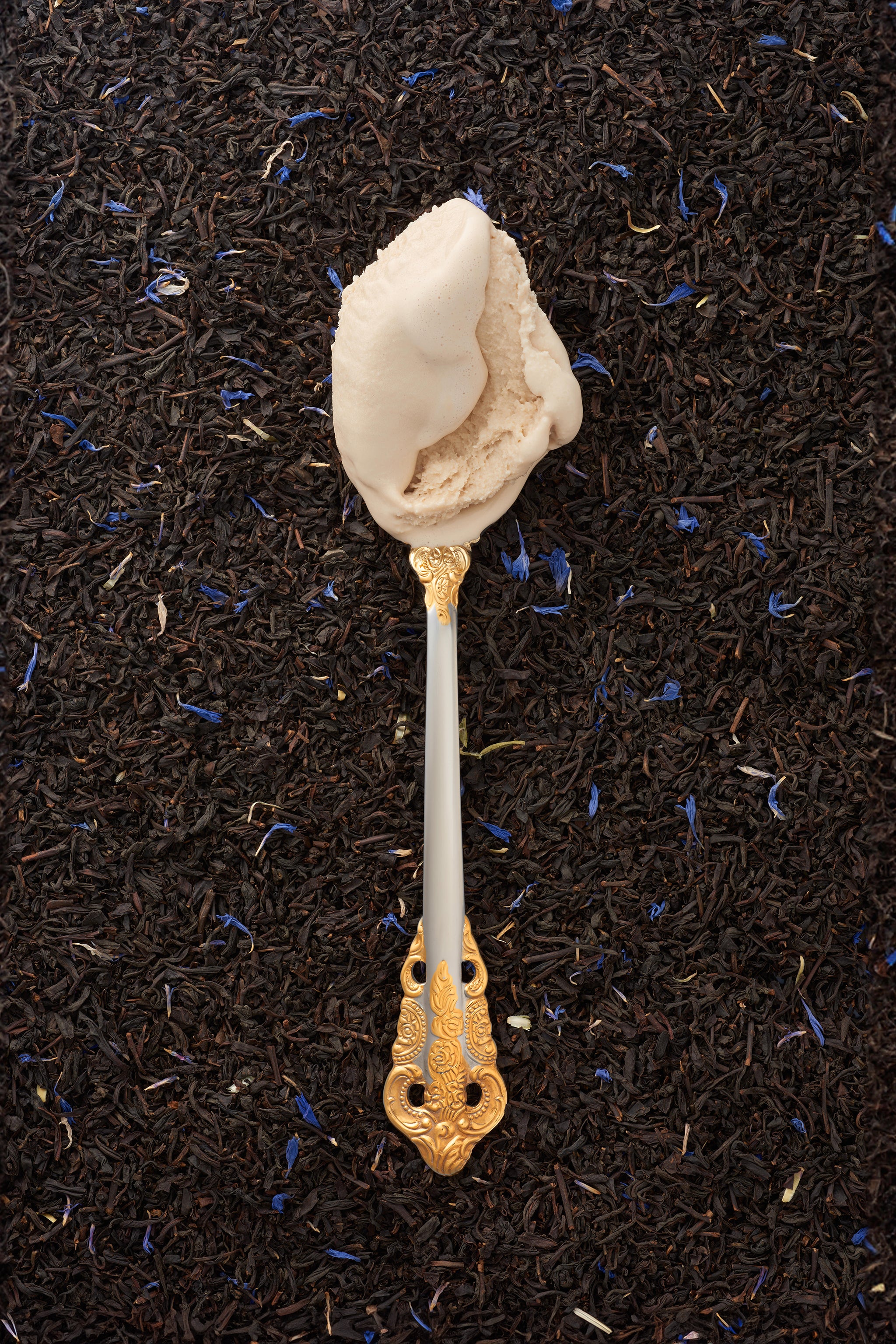 A scoop of earl grey kulfi in a silver spoon with a gold handle, with a backdrop of loose earl grey tea leaves.