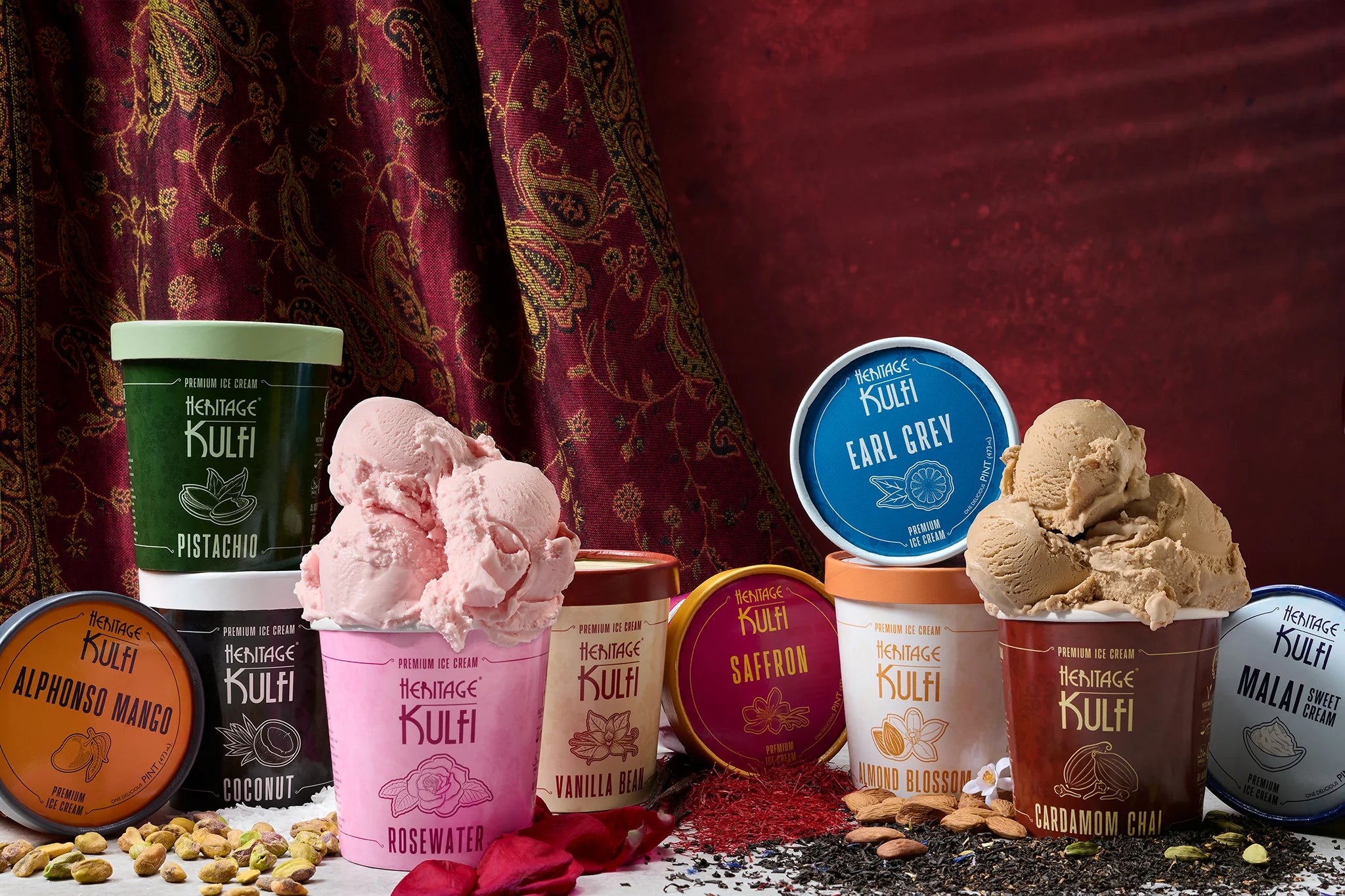 A group of all of the heritage kulfi flavors scattered on a table with their respective flavor elements sprinkled alongside, and a rich vibrant red tapestry in the background