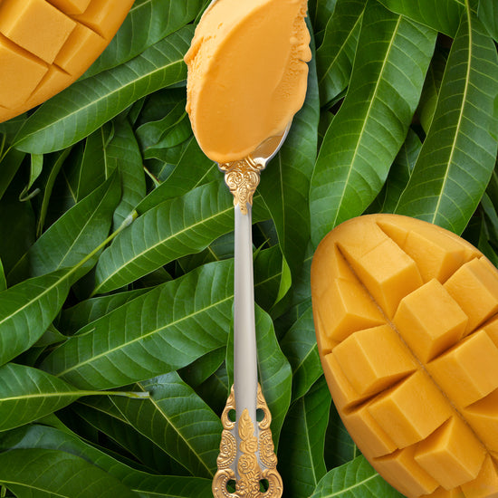 vibrant scoop of mango kulfi in a silver spoon with a gold handle, set against a backdrop of sliced mangoes and green leaves.