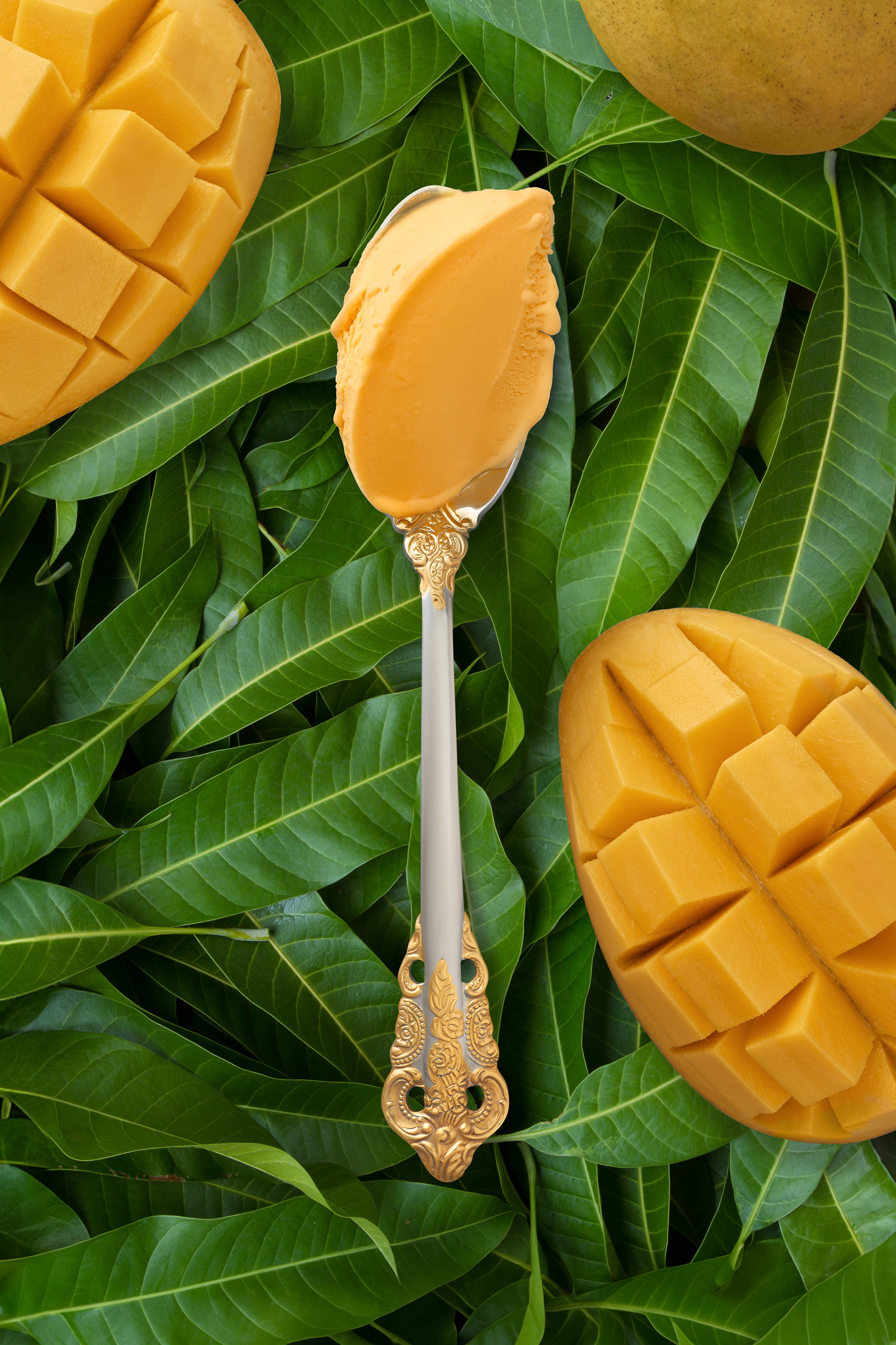  vibrant scoop of mango kulfi in a silver spoon with a gold handle, set against a backdrop of sliced mangoes and green leaves.