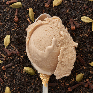 A closeup of chai colored icecream on a silver spoon, with a backdrop of chai tea leaves and cardamom pods