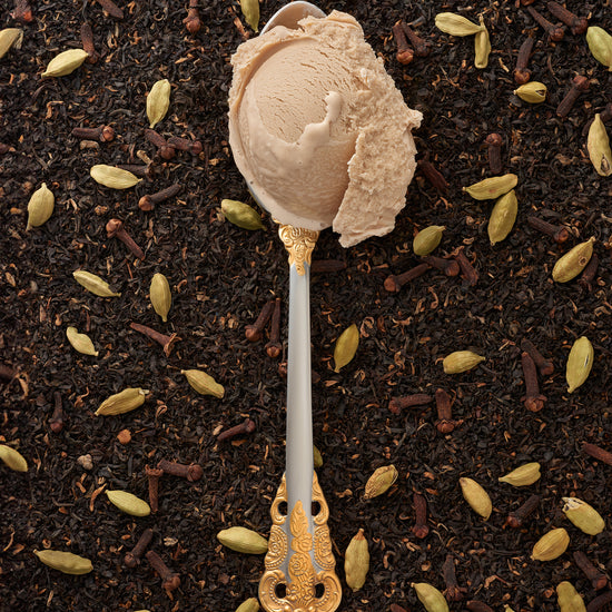 A scoop of cardamom-chai kulfi in a silver spoon with a gold handle, with a scattering of chai tea and cardamom pods in the background.