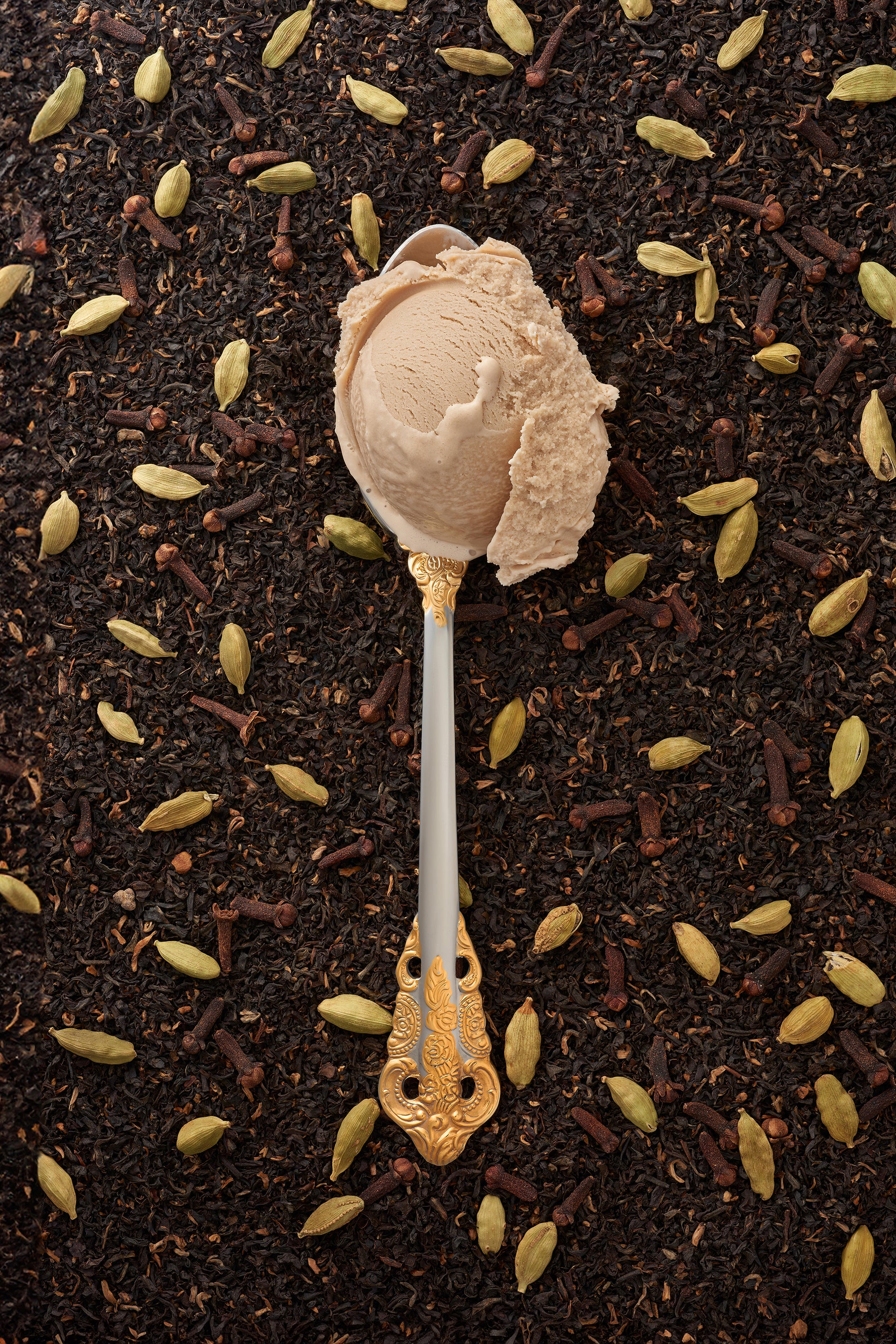 A scoop of cardamom-chai kulfi in a silver spoon with a gold handle, with a scattering of chai tea and cardamom pods in the background.