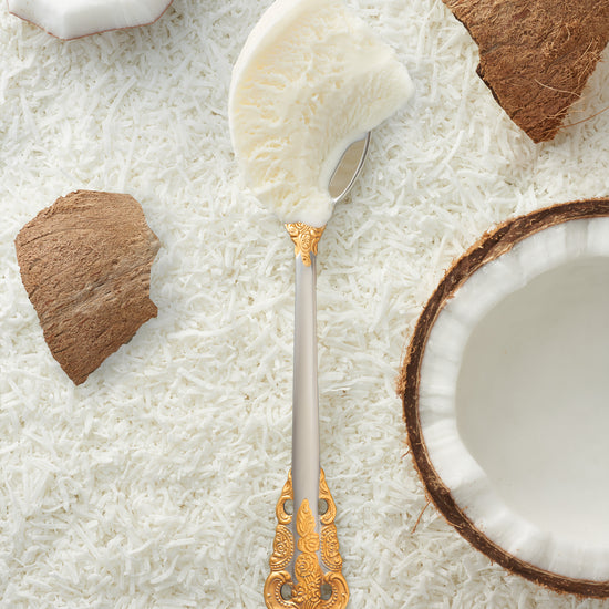 A scoop of coconut kulfi in a silver spoon with a gold handle, surrounded by pieces of fresh coconut.