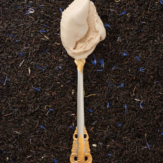 A scoop of earl grey kulfi in a silver spoon with a gold handle, with a backdrop of loose earl grey tea leaves.