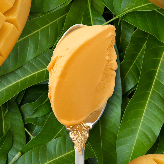 A closeup of a bright colored scoop of mango kulfi on a silver spoon, against a backdrop of mango leaves