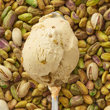 A closeup of a scoop of pistachio kulfi in a silver spoon, surrounded by a scattering of pistachios.