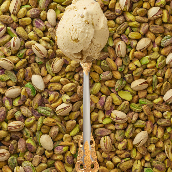 A scoop of pistachio kulfi in a silver spoon with a gold handle, surrounded by a scattering of pistachios.