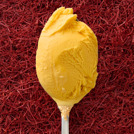 A closeup of a scoop of saffron kulfi in a silver spoon with a gold handle, with a background entirely covered in saffron threads