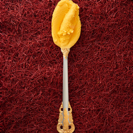 A scoop of saffron kulfi in a silver spoon with a gold handle, with a background entirely covered in saffron threads