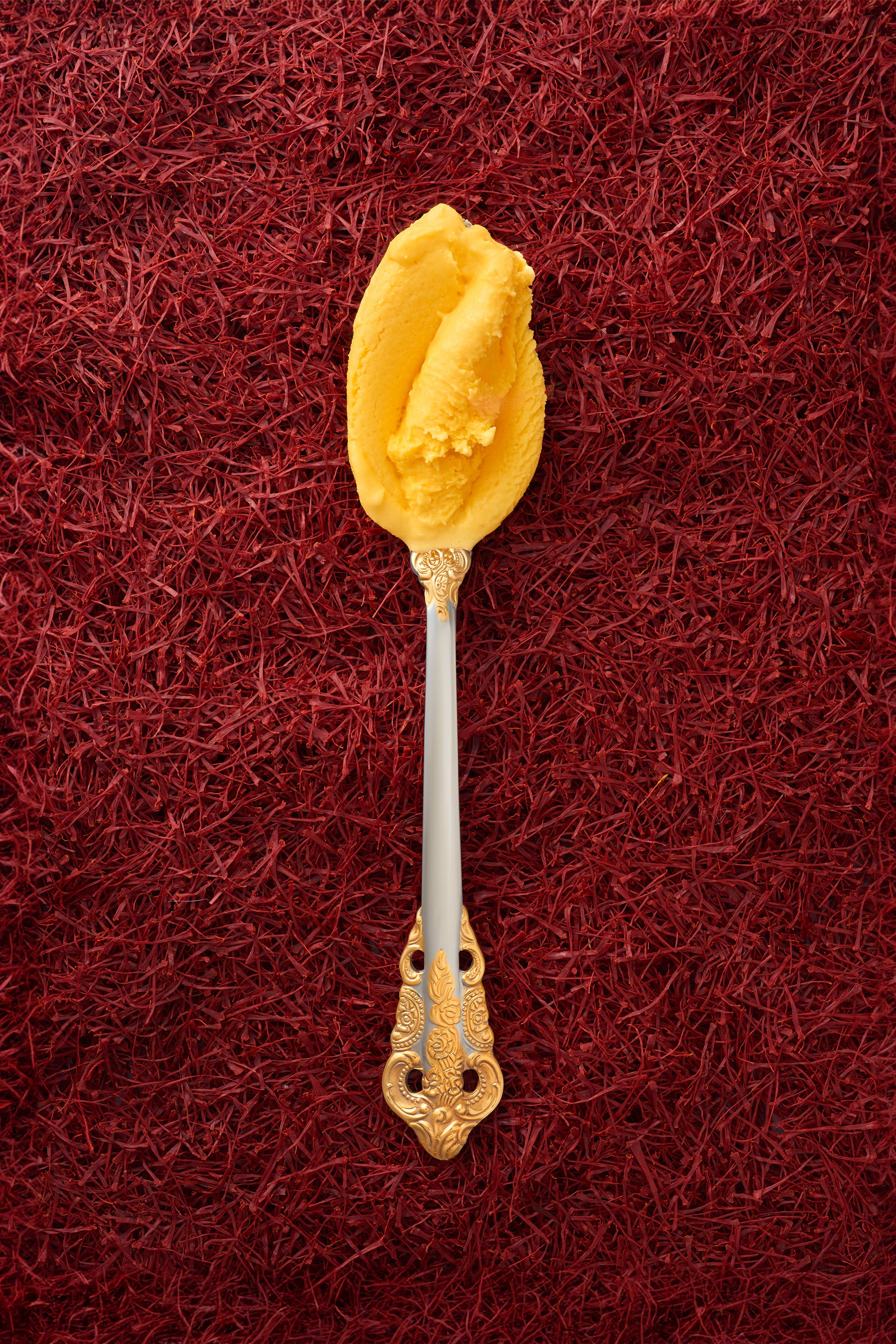 A scoop of saffron kulfi in a silver spoon with a gold handle, with a background entirely covered in saffron threads