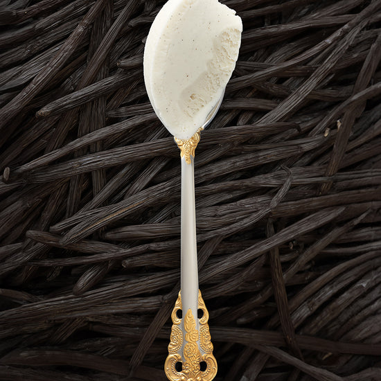 A scoop of vanilla bean kulfi in a silver spoon with a gold handle, with vanilla beans covering the background completely.