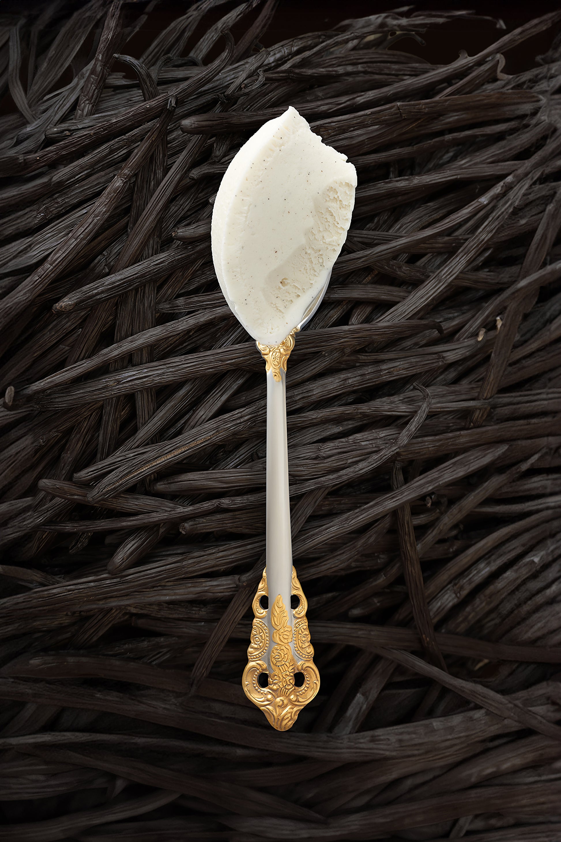 A scoop of vanilla bean kulfi in a silver spoon with a gold handle, with vanilla beans covering the background completely.