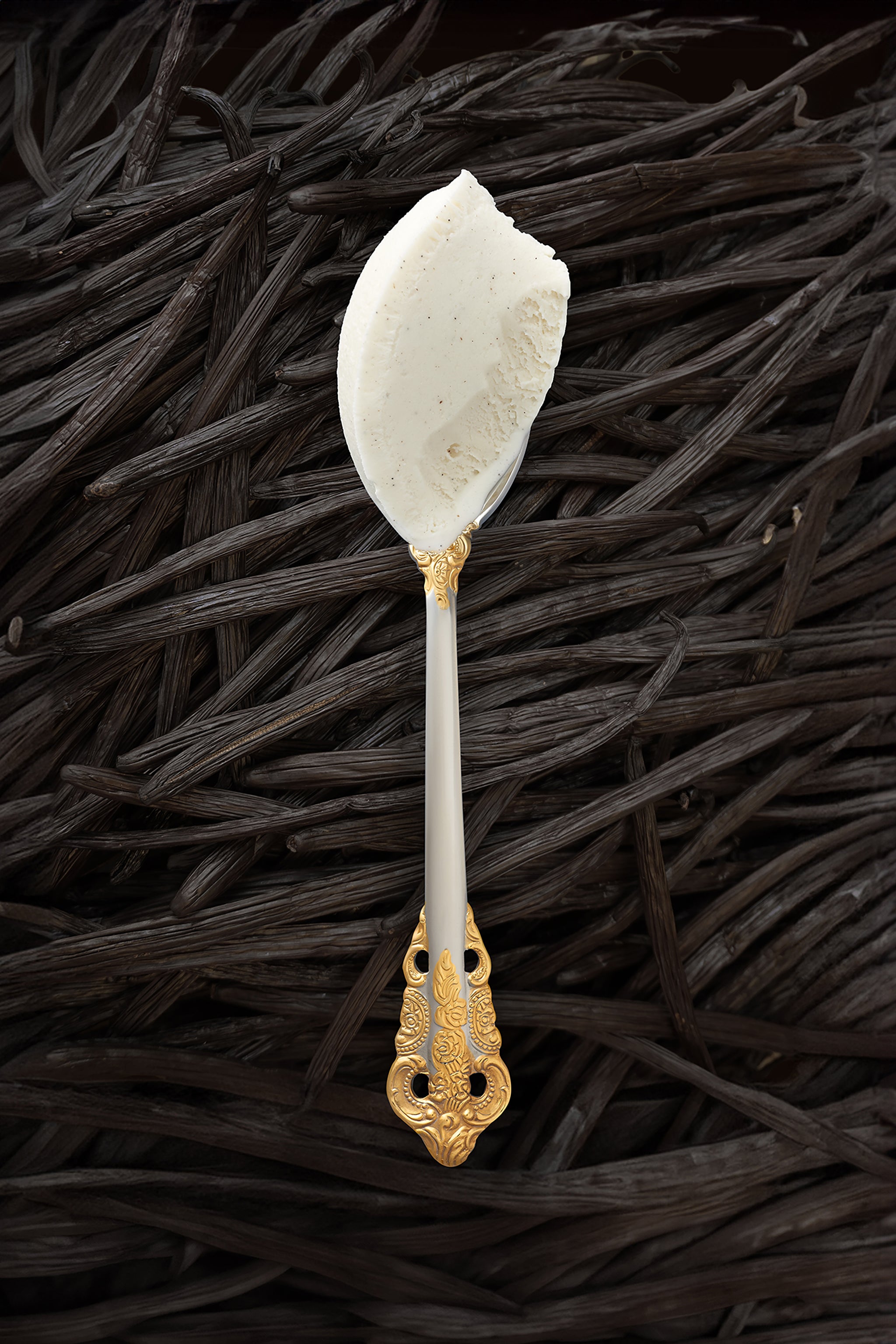 A scoop of vanilla bean kulfi in a silver spoon with a gold handle, with vanilla beans covering the background completely.